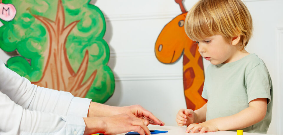 Boy with adult in kindergarten put figures on the table Little blond boy with help of adult in kindergarten put figures made of geometric shapes on the table